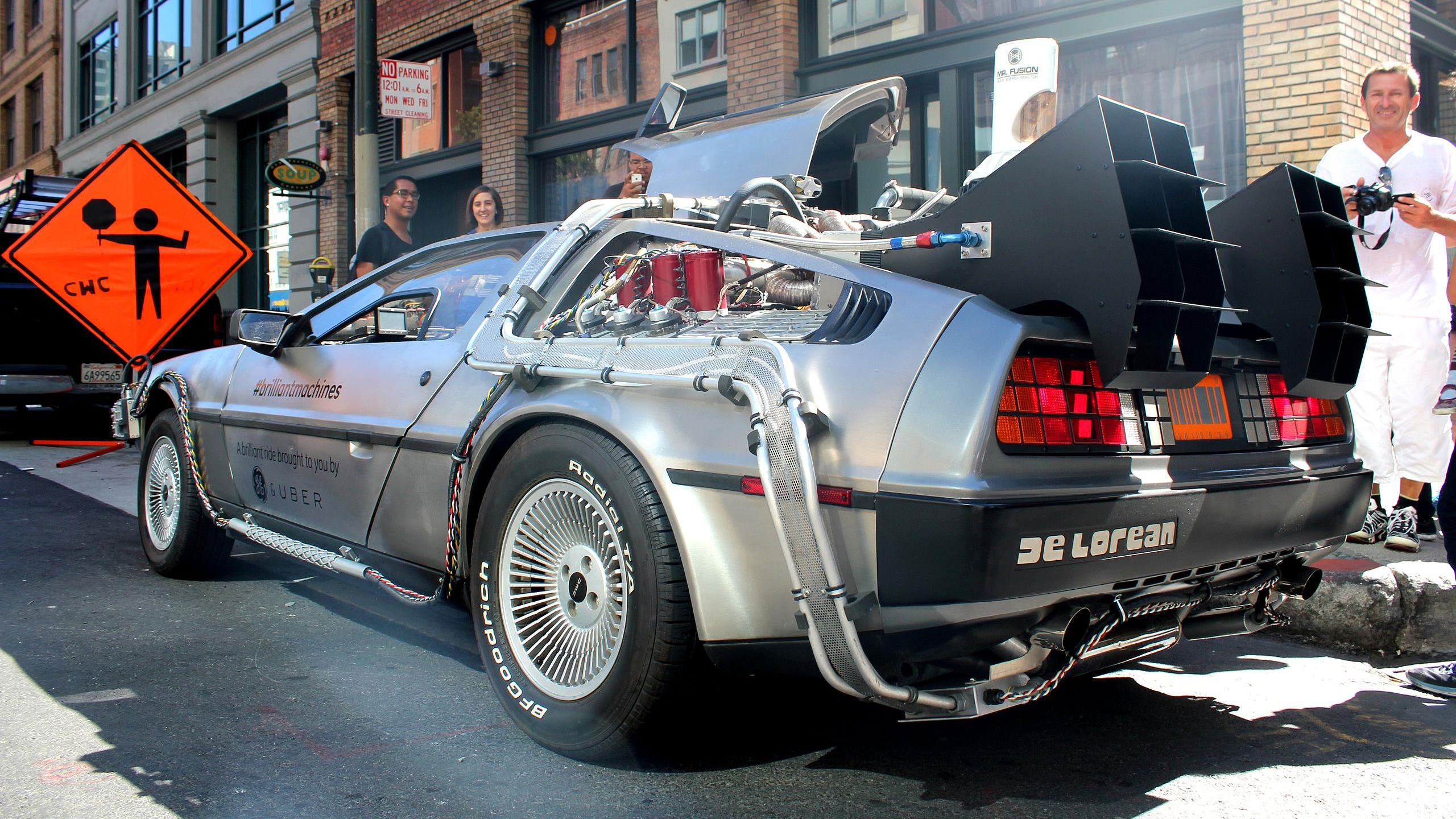 A silver DeLorean car, modified to resemble the time machine from "Back to the Future," is parked on a street. People are standing nearby, marveling at the iconic vehicle, while an orange construction sign looms in the background, hinting at disruptions in travel time physics.
