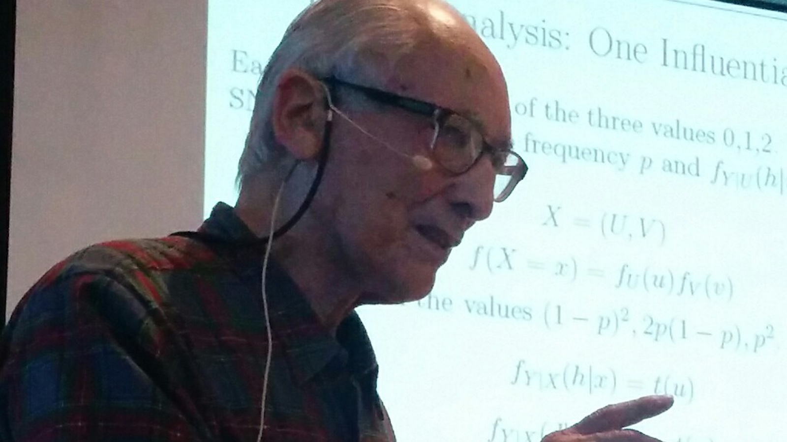 An elderly man speaks while wearing a microphone headset, with a mathematical presentation projected behind him.