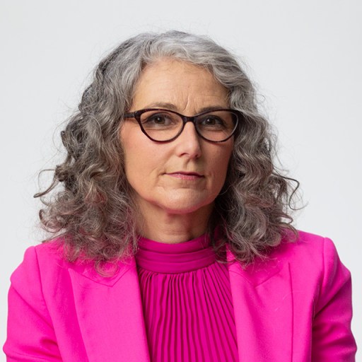 A gray-haired woman wearing glasses and a bright pink blazer with a pleated blouse, looking at the camera against a plain white background.