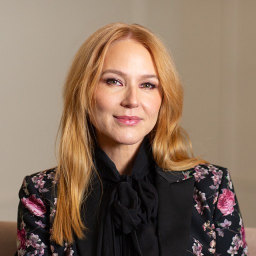 A person with long reddish-blonde hair, wearing a black floral-patterned blazer, sits and smiles at the camera.