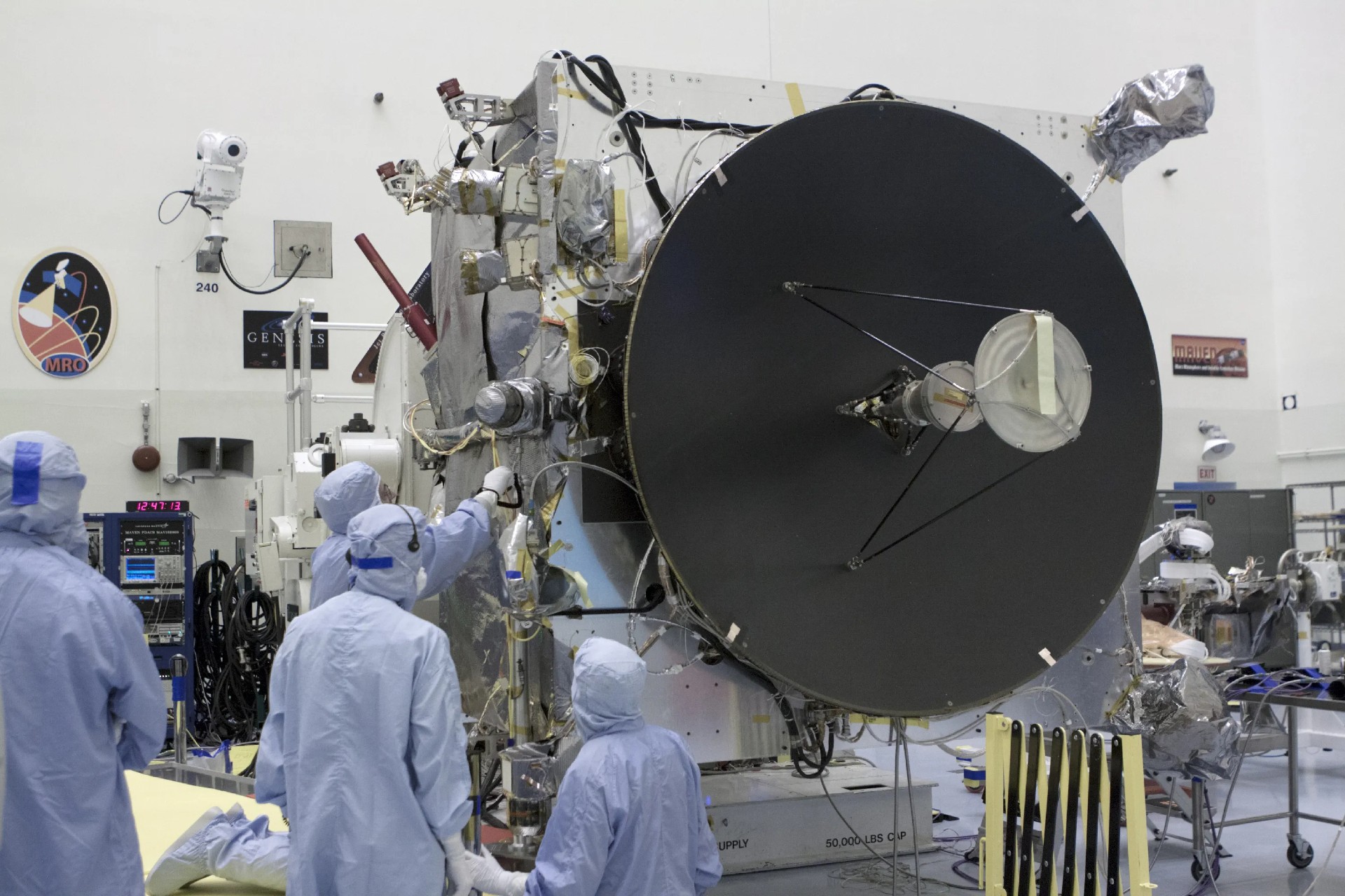 Scientists in blue protective suits work on a large satellite or spacecraft with a prominent circular dish in a laboratory setting.