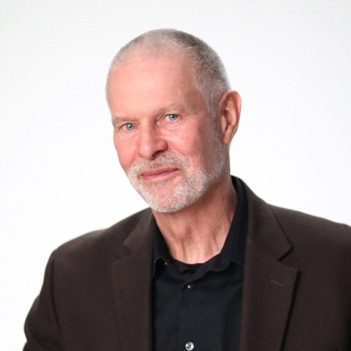 A man with short gray hair and a beard is wearing a dark blazer and black shirt, posing against a plain white background.