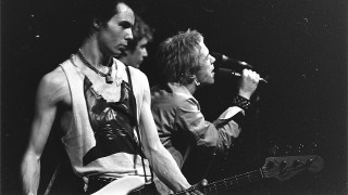 Black and white image of a band performing on stage. One musician is playing a bass guitar while another sings into a microphone. The scene, embodying punk-inspired leaders, has a dark background.