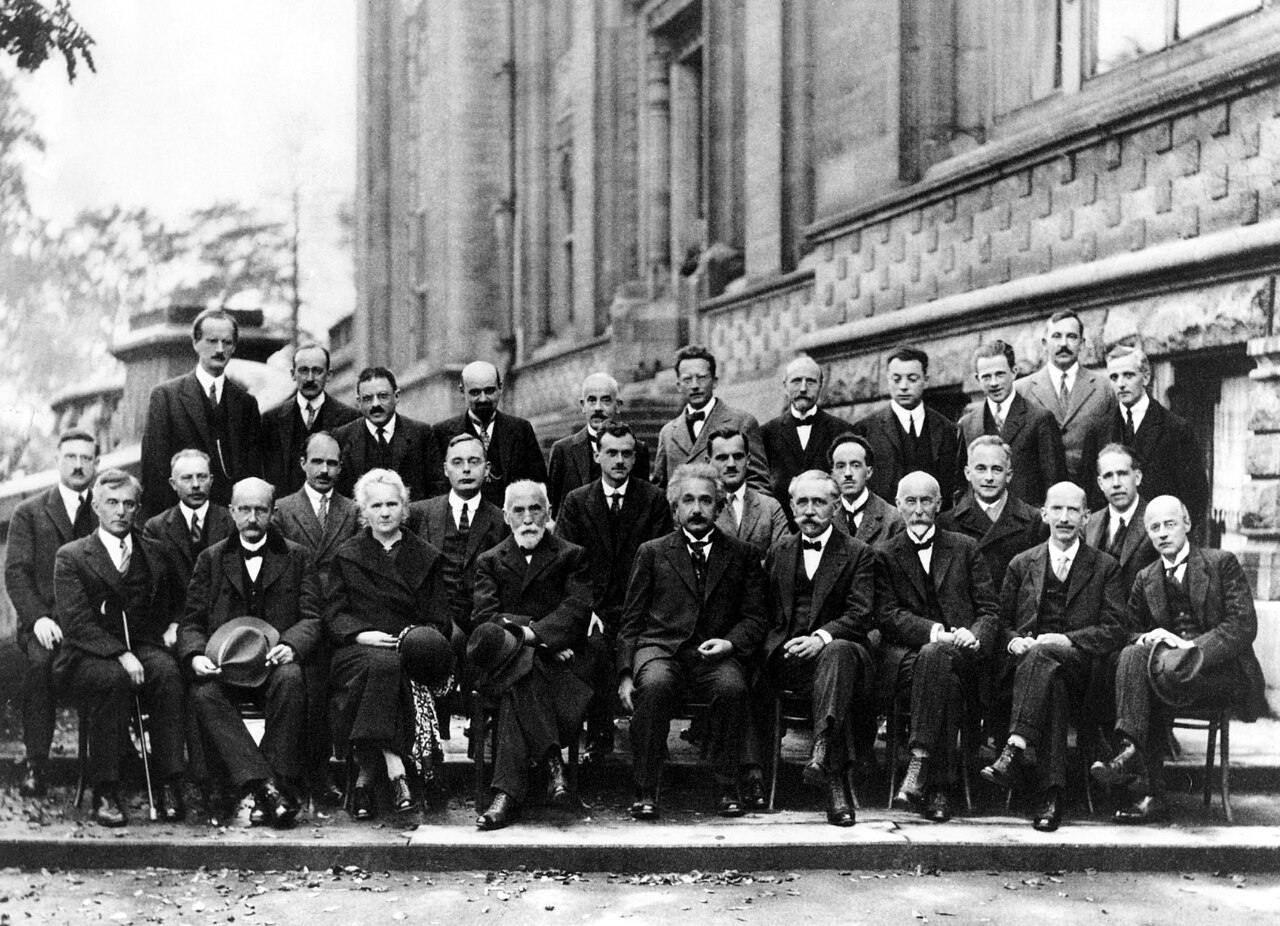 A group of approximately thirty formally dressed individuals, including men and a few women, are seated and standing in rows outside a building for a formal photograph.