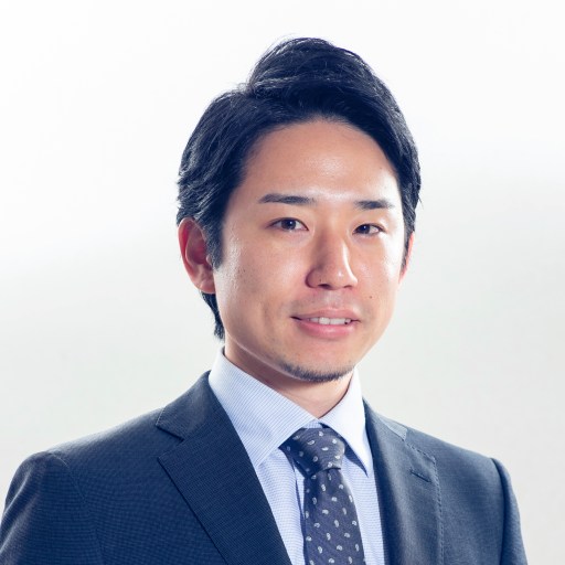 A man with short black hair is wearing a suit and tie. He has a slight smile and is looking straight at the camera against a plain background.