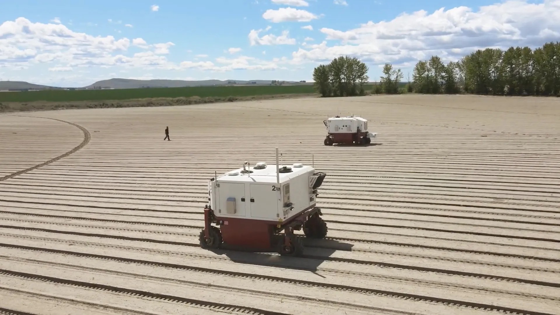 Two automated agricultural vehicles working in a large, flat, evenly plowed field with a person walking in the background. Sparse tree line and hill visible under a partly cloudy sky.