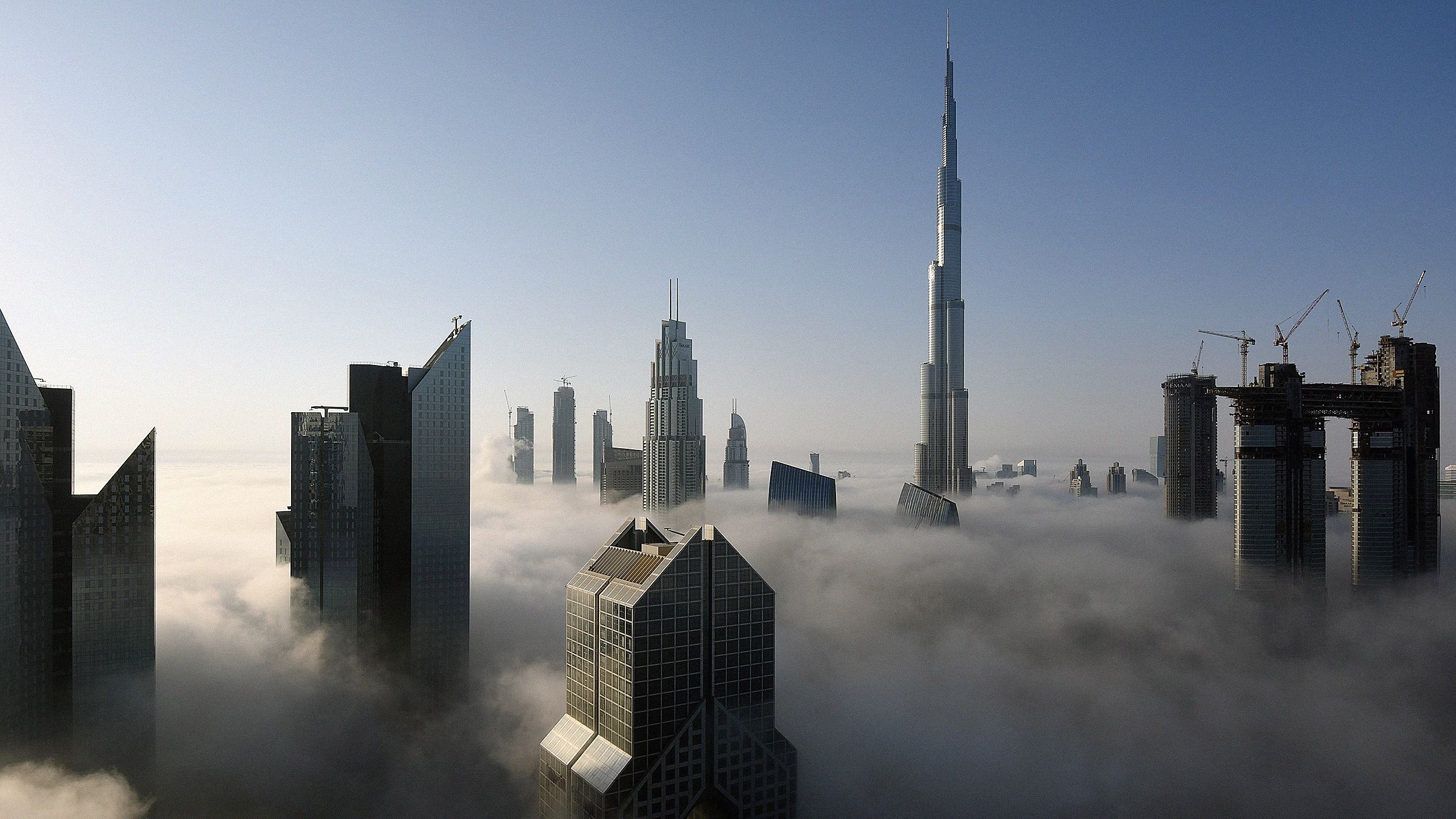 Skyline of a city with tall skyscrapers emerging above a thick layer of fog on a clear day, including one exceptionally tall building towering over the tallest buildings.