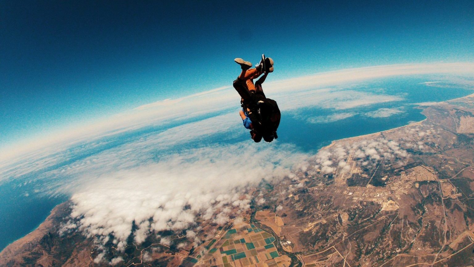 Two people are tandem skydiving, falling through the air above a landscape of fields, mountains, and coastline under a clear sky.