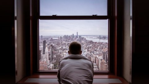 A person in a white shirt looks out a large window at a cityscape with skyscrapers and distant water under a cloudy sky.