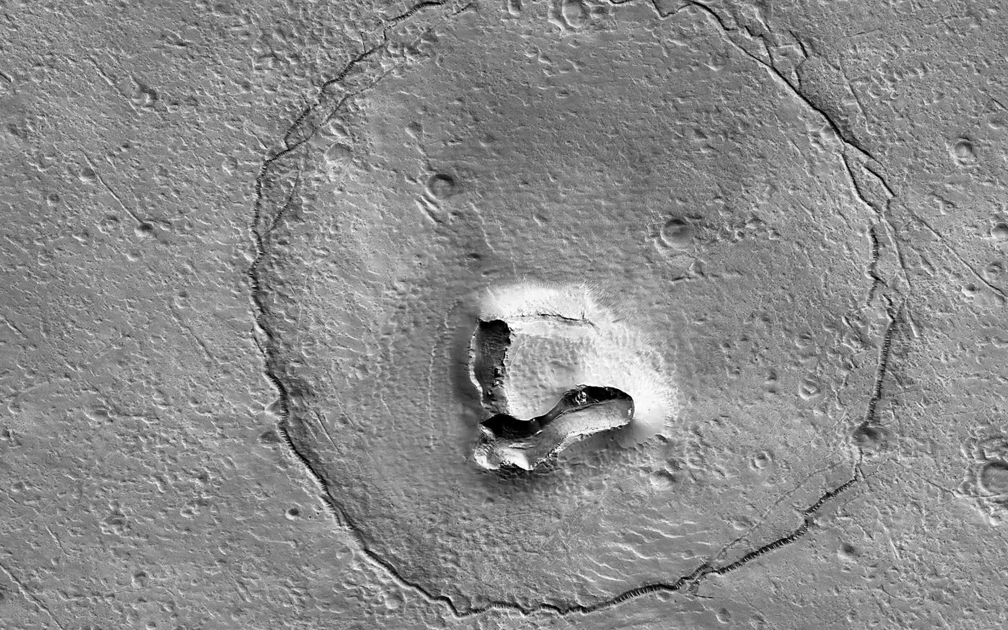 Satellite image of a large circular feature on a gray, rocky surface with a raised, irregularly shaped structure in the center and several small craters scattered around.