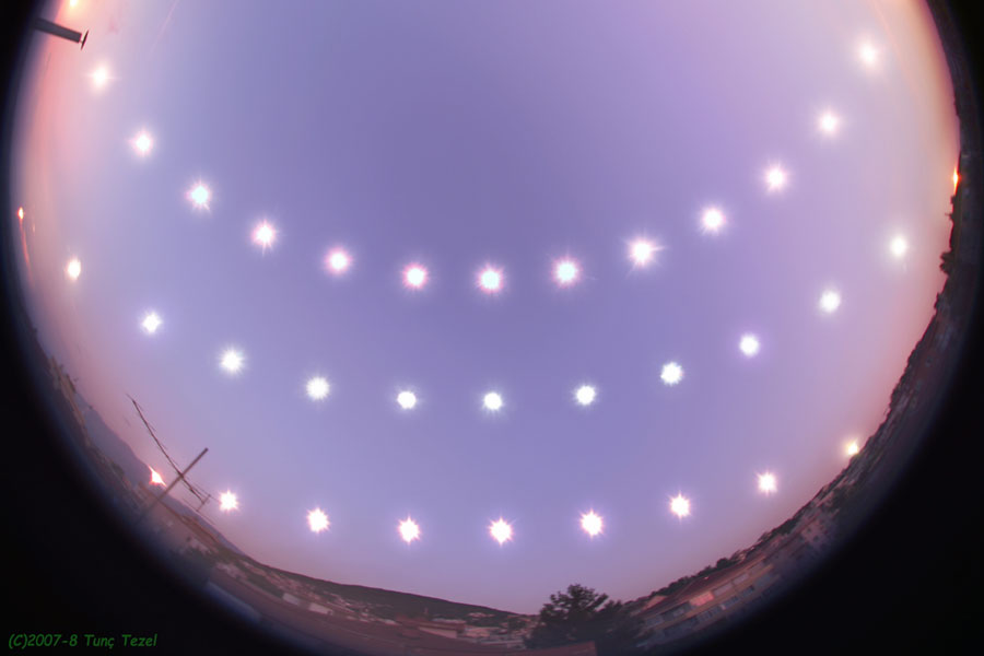A time-lapse photo showing the Sun's path over several days, arranged to form a smiley face pattern against a twilight background. Taken around the period of the earliest solstice, ©2007-8 Tunç Tezel is credited in the bottom left corner.