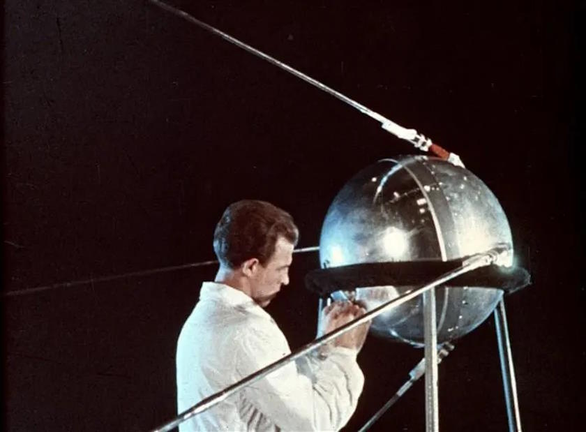A man in a white coat inspects the spherical satellite, Sputnik 1, which is mounted on a stand and visible with long antennas extending from its body. As he examines the iconic spacecraft, one can't help but think of Bill Anders capturing Earth from Apollo 8.