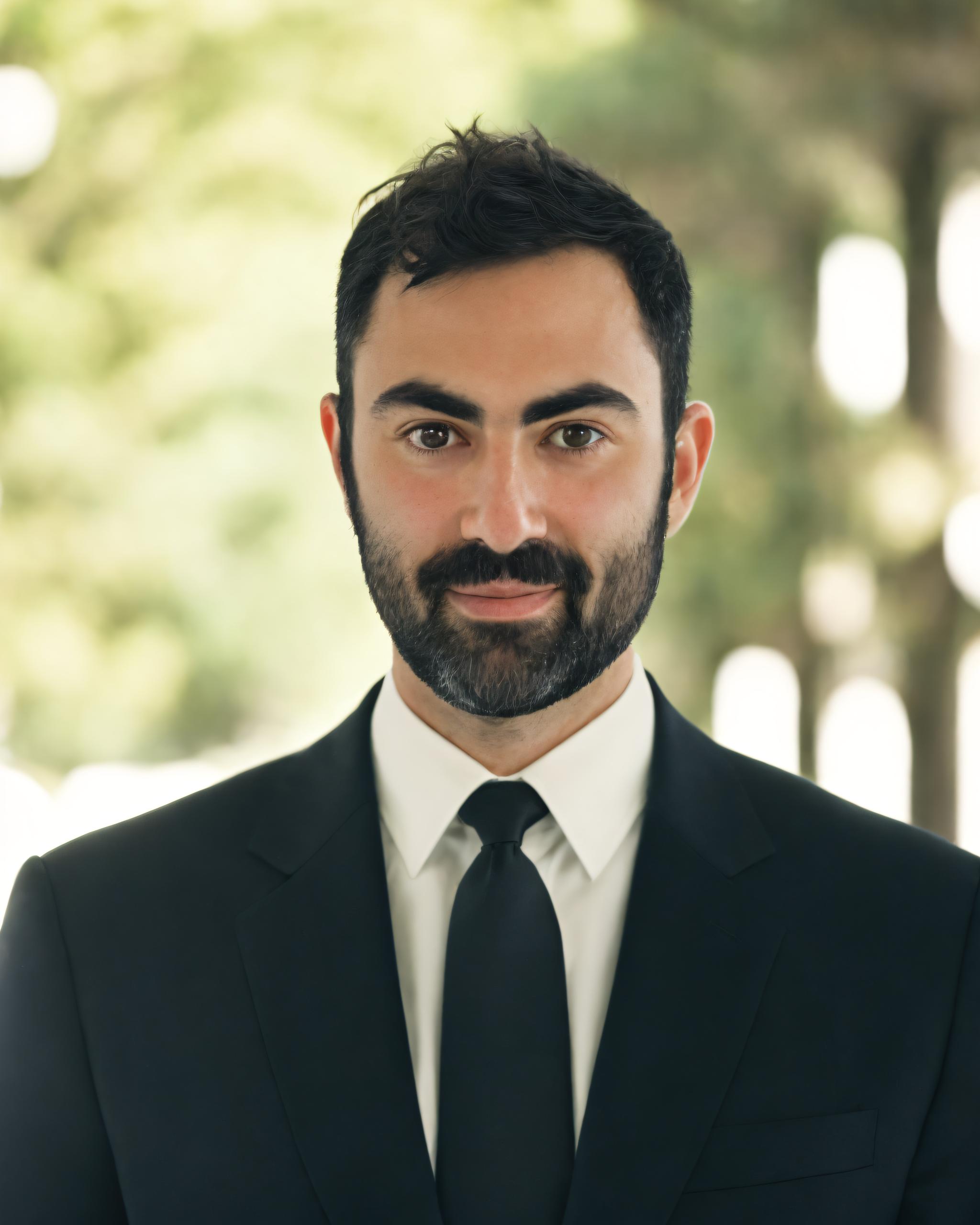 A bearded man in a black suit and tie stands outdoors with blurred green foliage in the background.