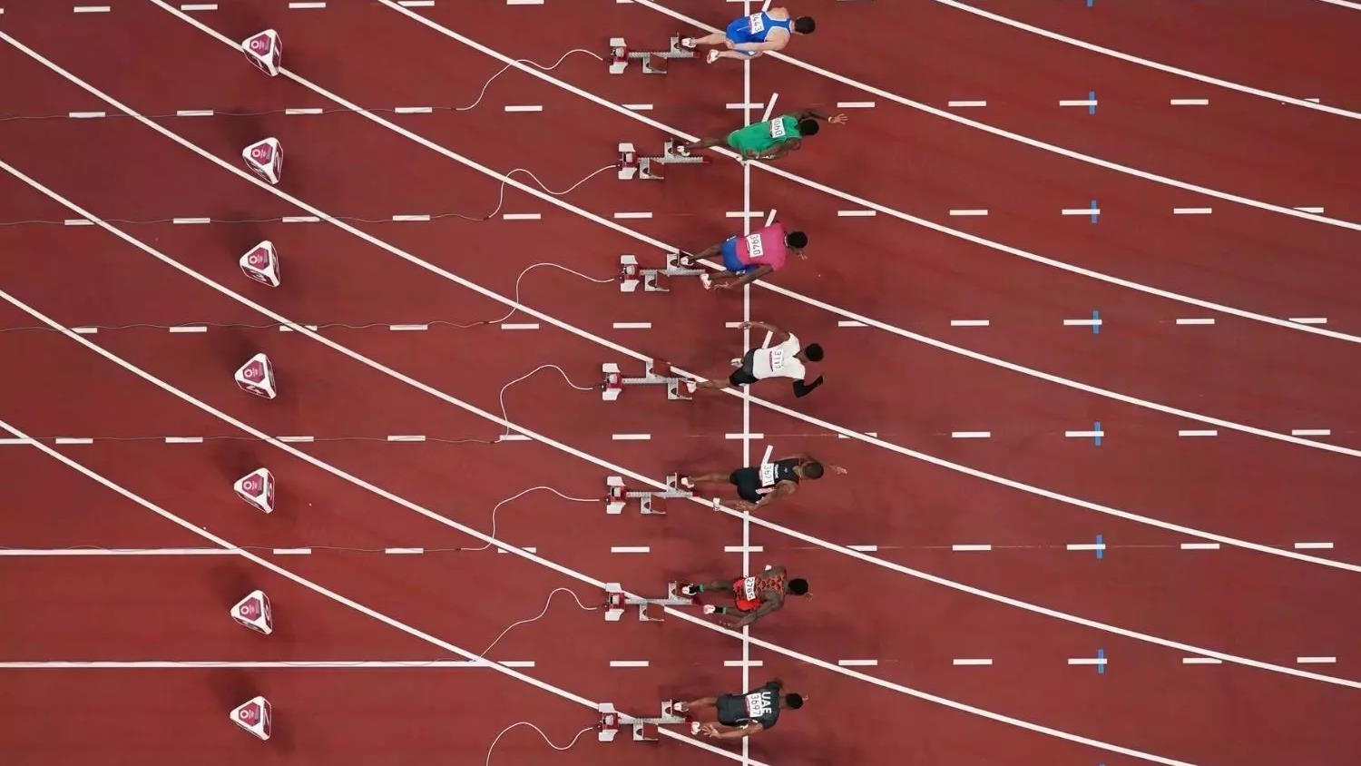 Overhead view of athletes in starting blocks on a track, preparing for the fastest 100 meters. Marked lanes and starting lines are visible.