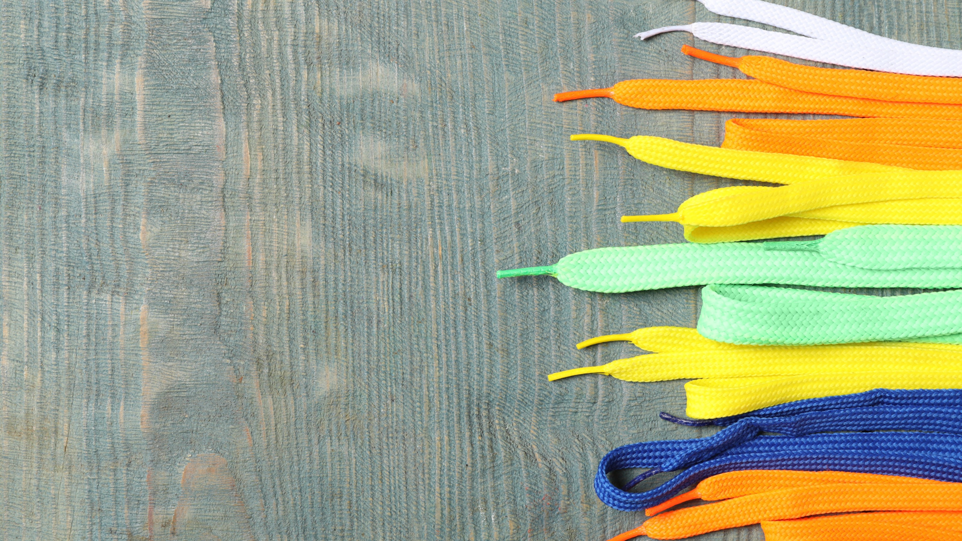 Multicolored shoelaces in varying shades of yellow, green, blue, and orange are arranged neatly on the right side of a wooden surface. The left side of the surface is empty.