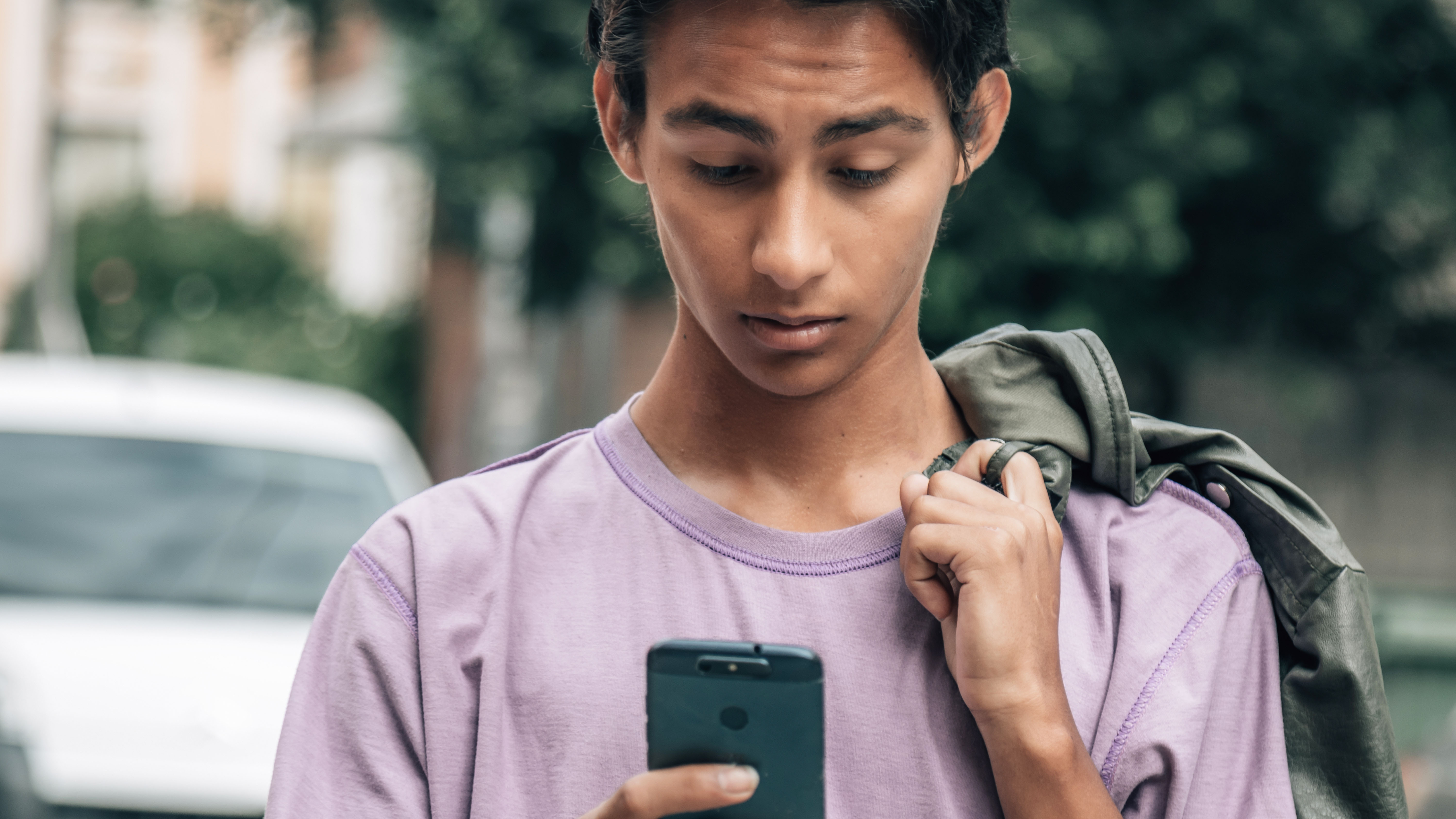 A young person in a purple shirt holds a phone, looking down at it with a jacket draped over one shoulder while standing outdoors, unaware of the potential dangers such as inadvertently accessing pornography on the internet.