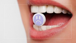 A close-up of a person's open mouth holding a pill between their teeth. The pill has a purple, sad face icon printed on it.