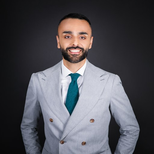 A man with a trimmed beard and dark hair is smiling and wearing a light gray double-breasted suit with a teal tie, standing against a plain dark background.