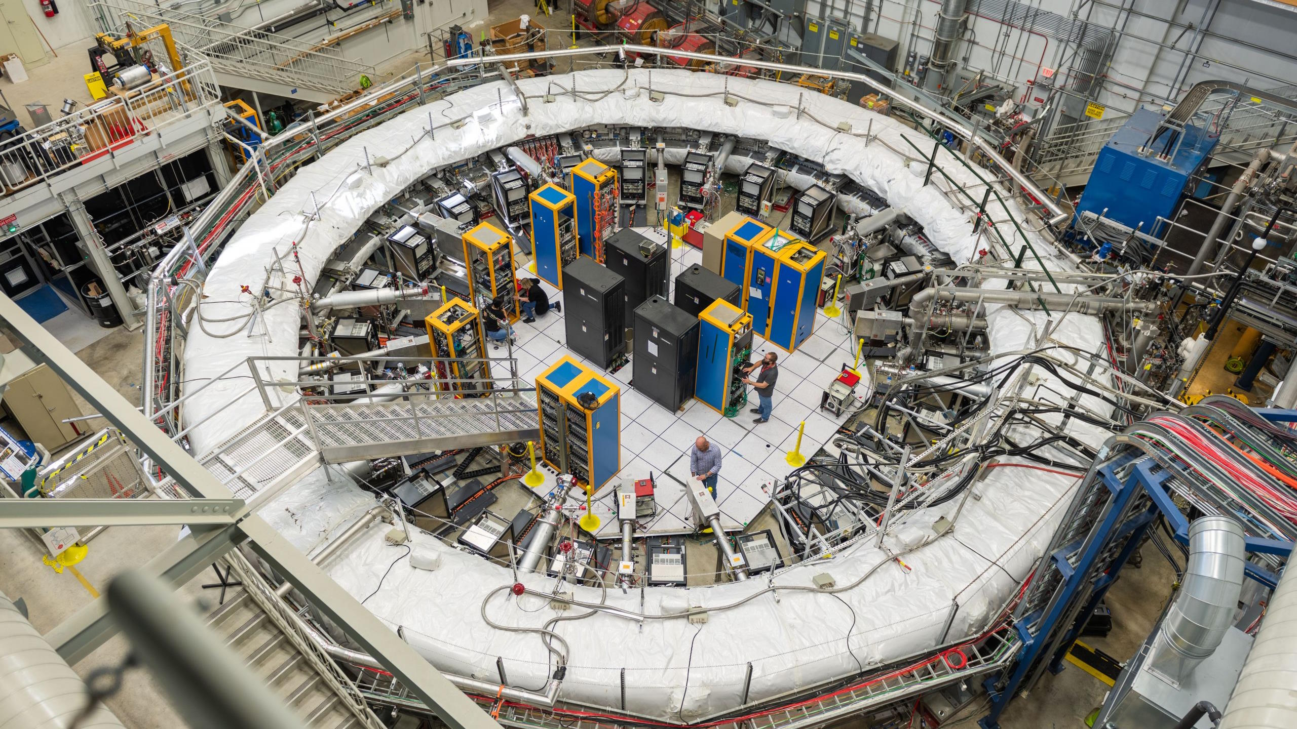 A large circular particle accelerator with several cables and machines is where engineers work inside and around the structure. The facility, dedicated to solving the muon g-2 anomaly, has platforms and specialized equipment surrounding the central structure.