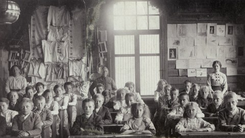 A black-and-white photo of young children sitting in a classroom. Two teachers stand at the back. Drawings and crafts are displayed on the walls, suggesting it was from an earlier historical period, where every good kid learned under watchful eyes.