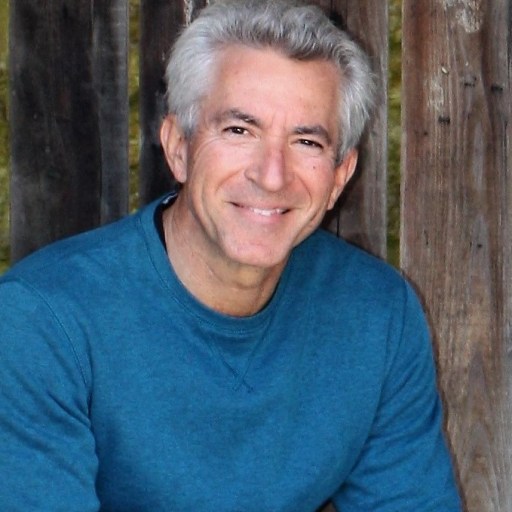 A man with gray hair wearing a blue sweater smiles while sitting in front of a wooden fence.