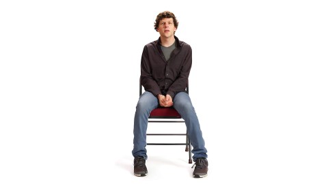 A person with curly hair, reminiscent of Jesse Eisenberg's style, wearing a dark long-sleeve shirt and jeans, sits on a chair against a plain white background.