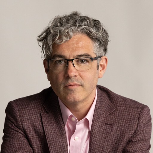 A man with curly gray hair and glasses wearing a maroon suit jacket and a pink shirt looks directly at the camera with a neutral expression.