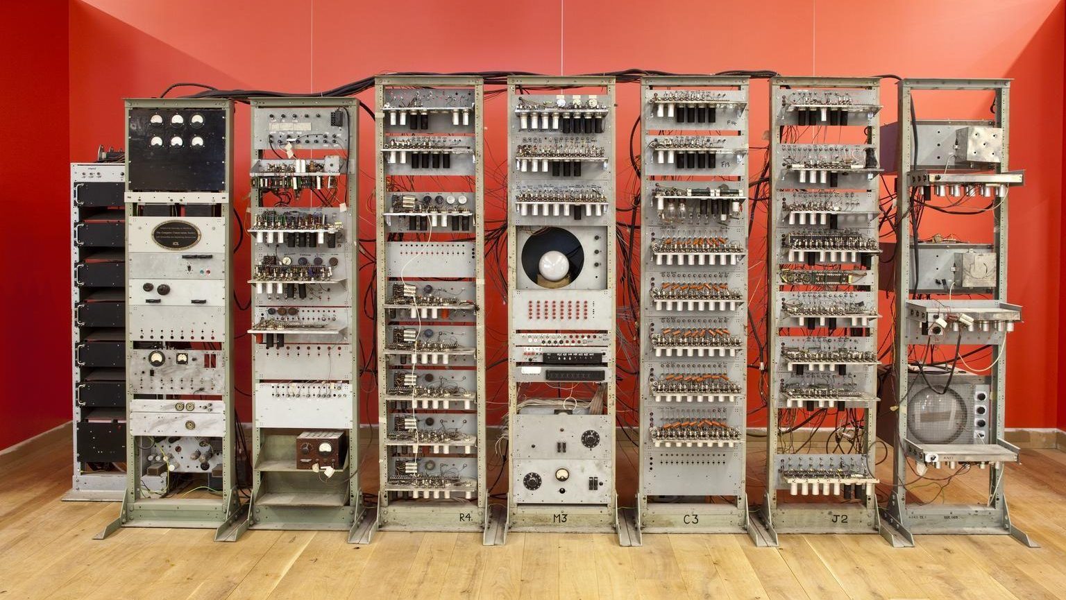 A vintage computer mainframe with multiple interconnected units, cables, and knobs, displayed in front of a red background on a wooden floor.
