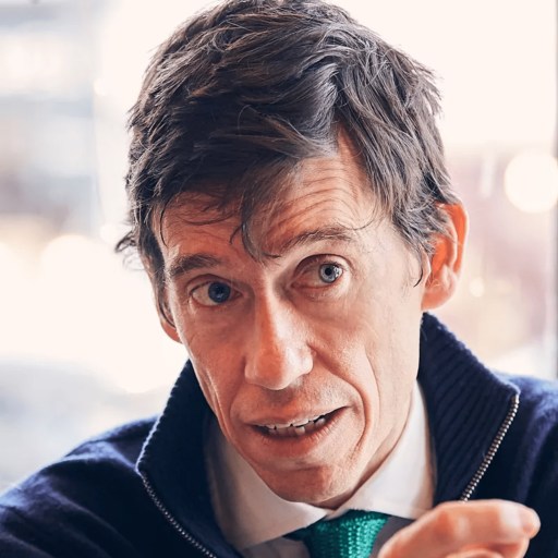 A man with short brown hair, dressed in a white shirt, blue jacket, and green tie, is captured in mid-conversation, looking slightly to the side against a blurred indoor background.