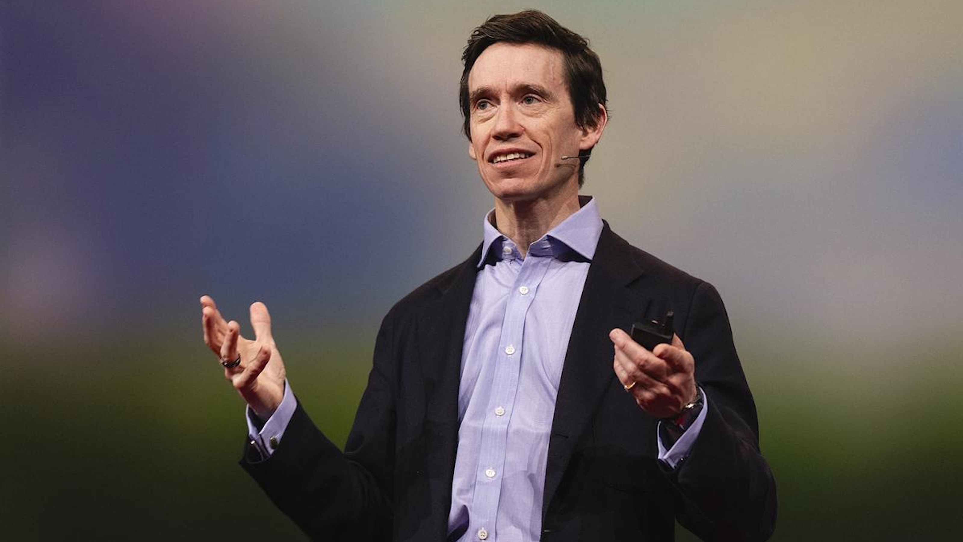 A person in a suit stands on stage, gesturing with both hands while speaking into a headset microphone, passionately discussing direct giving in their presentation.