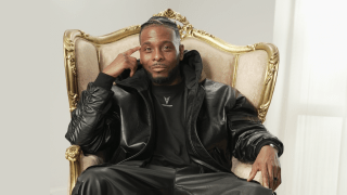 A man in a black leather outfit sits on an ornate, gold-trimmed chair, with one hand touching his temple and the other resting on the armrest.