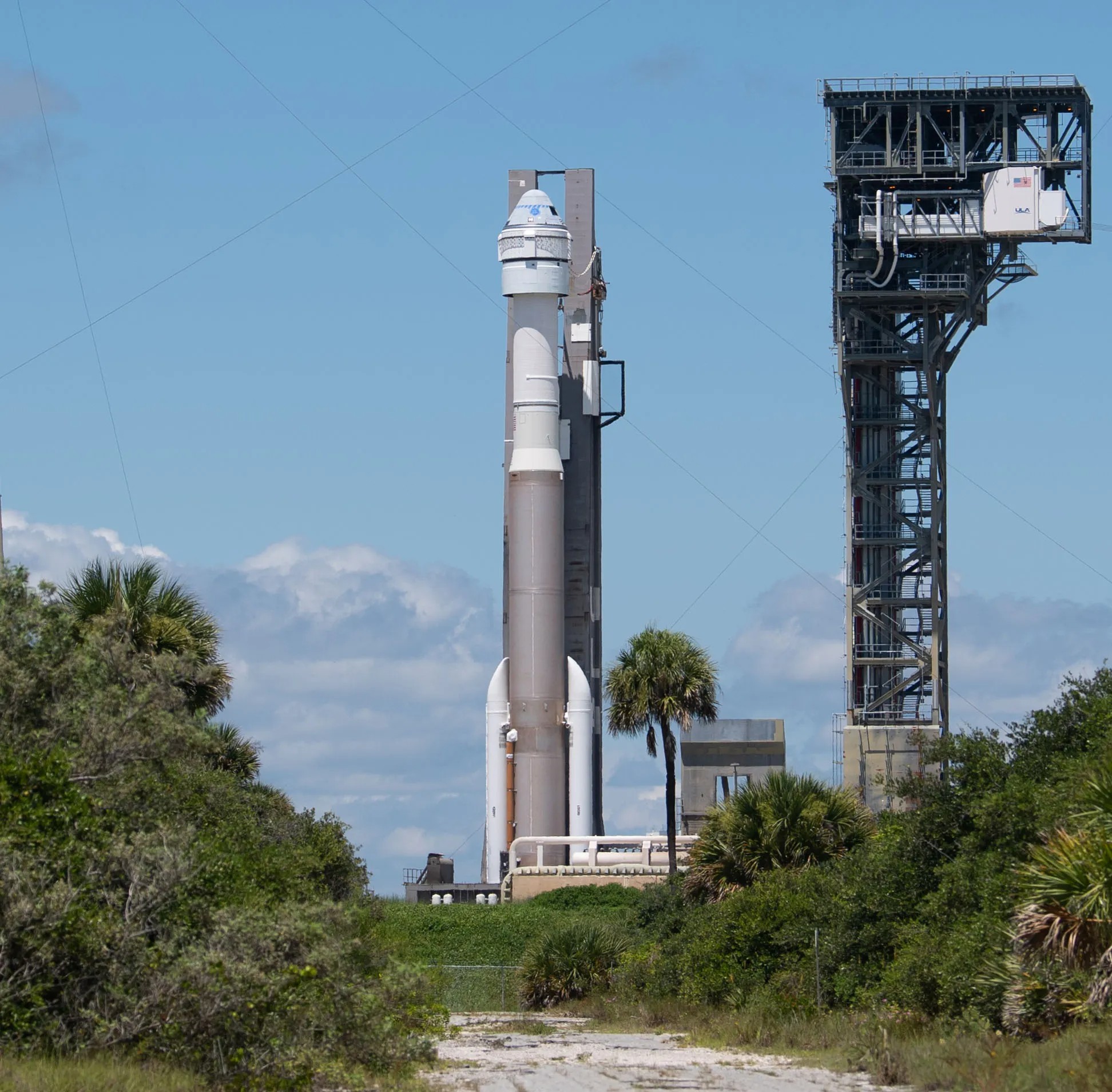 A rocket positioned vertically on a launch pad, surrounded by greenery and a blue sky with scattered clouds in the background.