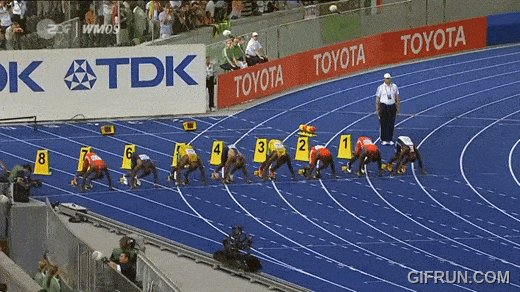 Athletes in starting positions on a blue track, ready for the fastest 100 meters race, with lanes numbered 1 to 8. A referee stands on the right near the starting line. Audience and equipment are visible in the background.