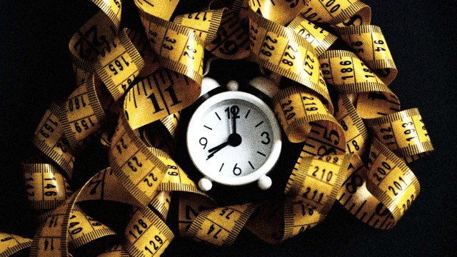 A small clock displaying 9:10 is surrounded by a tangled yellow measuring tape with black markings against a dark background, symbolizing the precision and commitment of intermittent fasting.