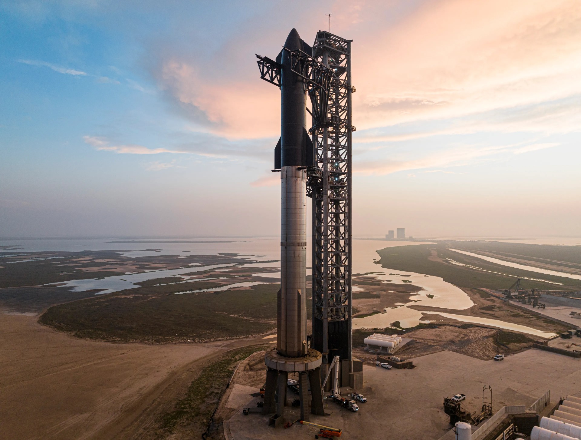 A large rocket stands vertically on a launch pad with support structures at sunset, surrounded by a vast landscape of wetlands and water channels.