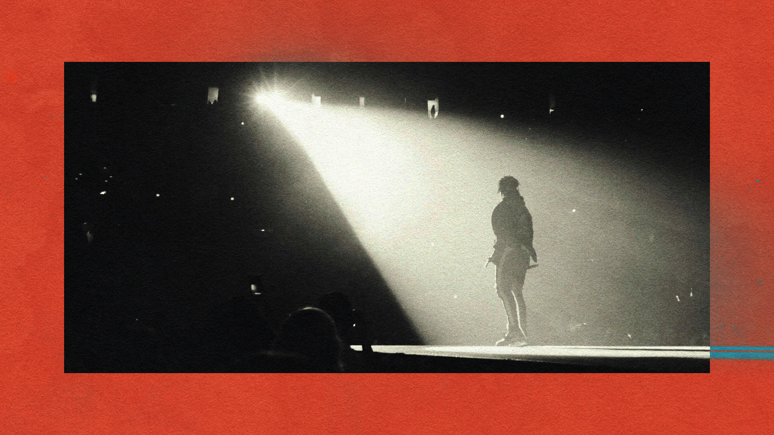 Black and white photo of a person on stage, the spotlight effect illuminating them from behind, casting dramatic shadows. A red border frames the image.