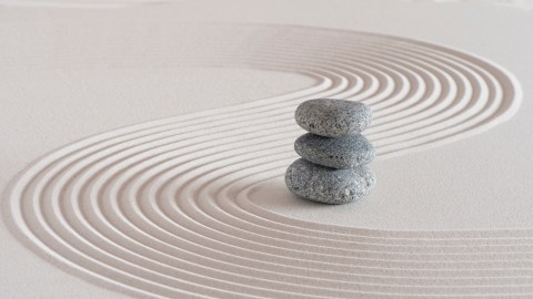 A stack of three grey pebbles is centered on a smooth sand surface with neatly raked curved lines forming a pattern around the stones.