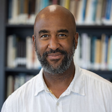 A person with a beard and bald head wearing a white shirt stands in front of shelves filled with books.