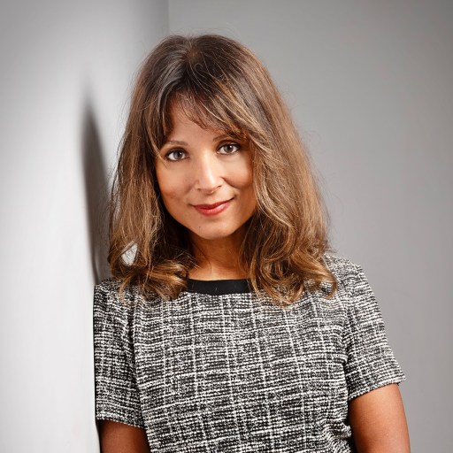 Person with shoulder-length brown hair and bangs, wearing a short-sleeve gray checkered dress, standing against a light gray wall.