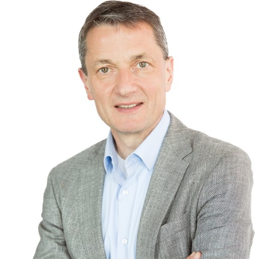 A man wearing a light gray blazer over a blue shirt stands with his arms crossed against a plain white background.