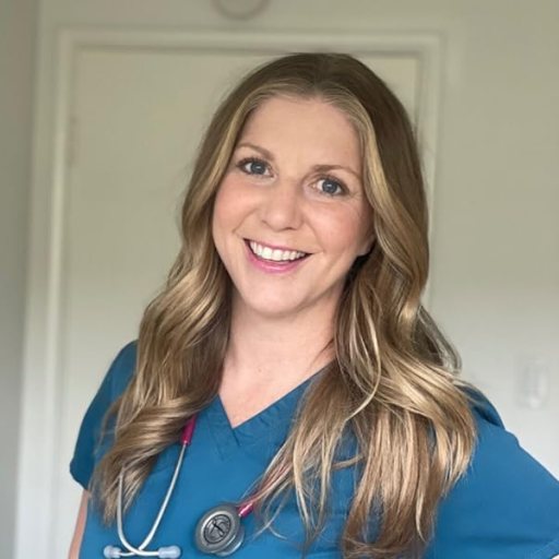 A person with long, wavy hair wearing blue scrubs and a stethoscope around their neck stands smiling in front of a plain white door.