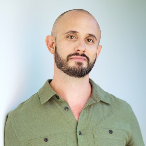 A man with a shaved head and a beard is wearing a green button-up shirt and standing against a plain background.