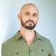 A man with a shaved head and a beard is wearing a green button-up shirt and standing against a plain background.