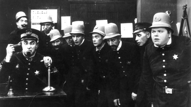 A group of vintage uniformed men, some wearing helmets, appear startled or curious while standing in what seems to be an office setting. The man on the left is speaking into a telephone, possibly exemplifying the Peter Principle as he manages the unexpected situation.