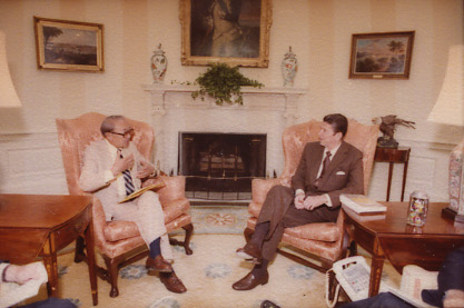 Two individuals are seated and engaged in conversation in a formal room with a fireplace. One is holding papers, and the other is sitting with legs crossed. Artwork and decor adorn the space.