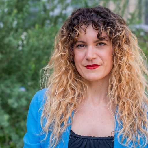 A person with curly hair and a blue blazer poses outdoors in front of greenery.