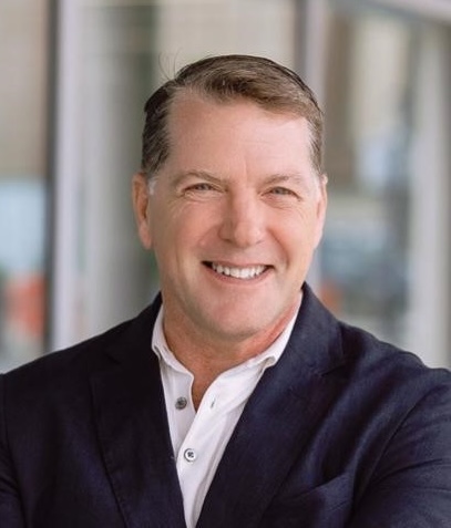 A man in a dark jacket and white shirt stands indoors, smiling at the camera.