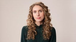 A person with long, curly blonde hair wearing a dark green blouse stands against a plain background.