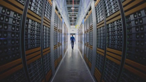 A wide shot of a man walking down an aisle between tall stacks of server racks in a data center filled with computer hardware.