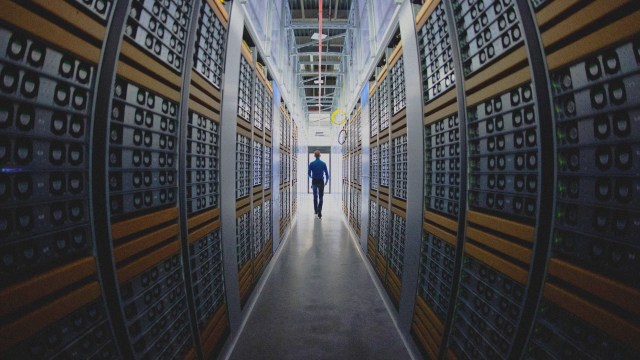 A wide shot of a man walking down an aisle between tall stacks of server racks in a data center filled with computer hardware.